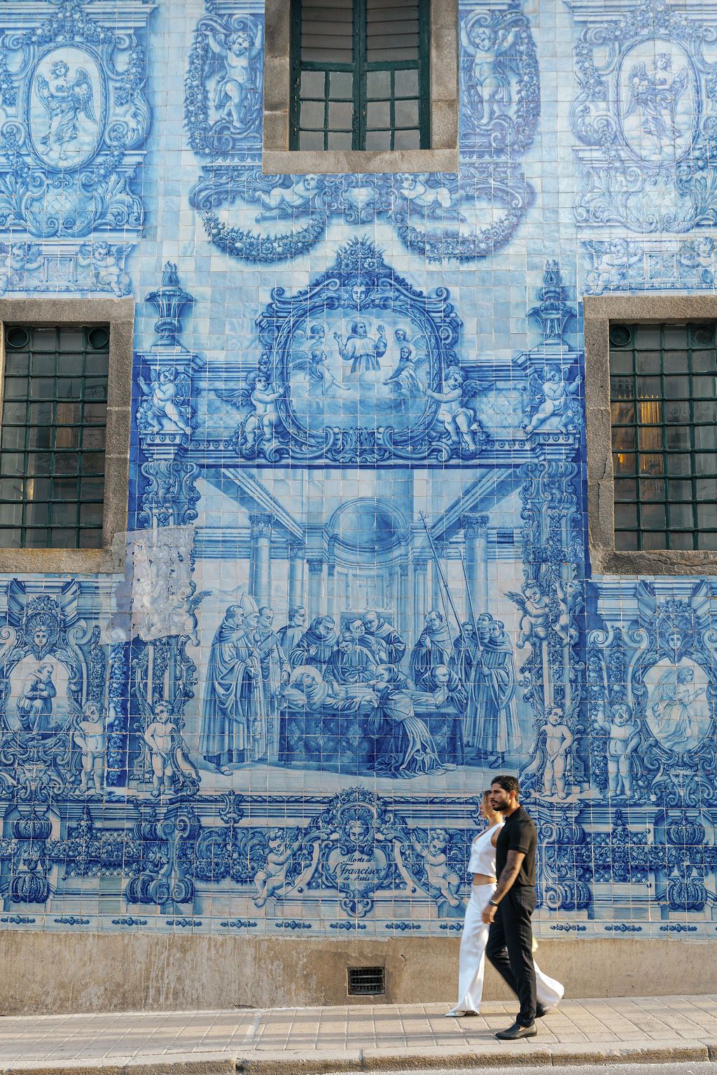 A couple poses against a blue and white tiled wall featuring traditional Portuguese azulejo artwork in Porto, Portugal.