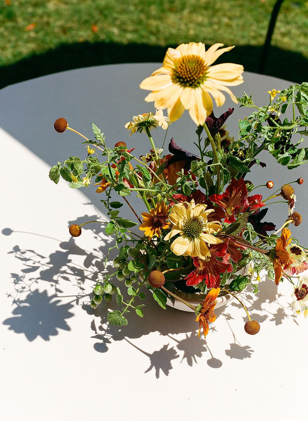 A bright yellow daisy flower arrangement with mixed foliage in a vase casting shadows on a white table surface.