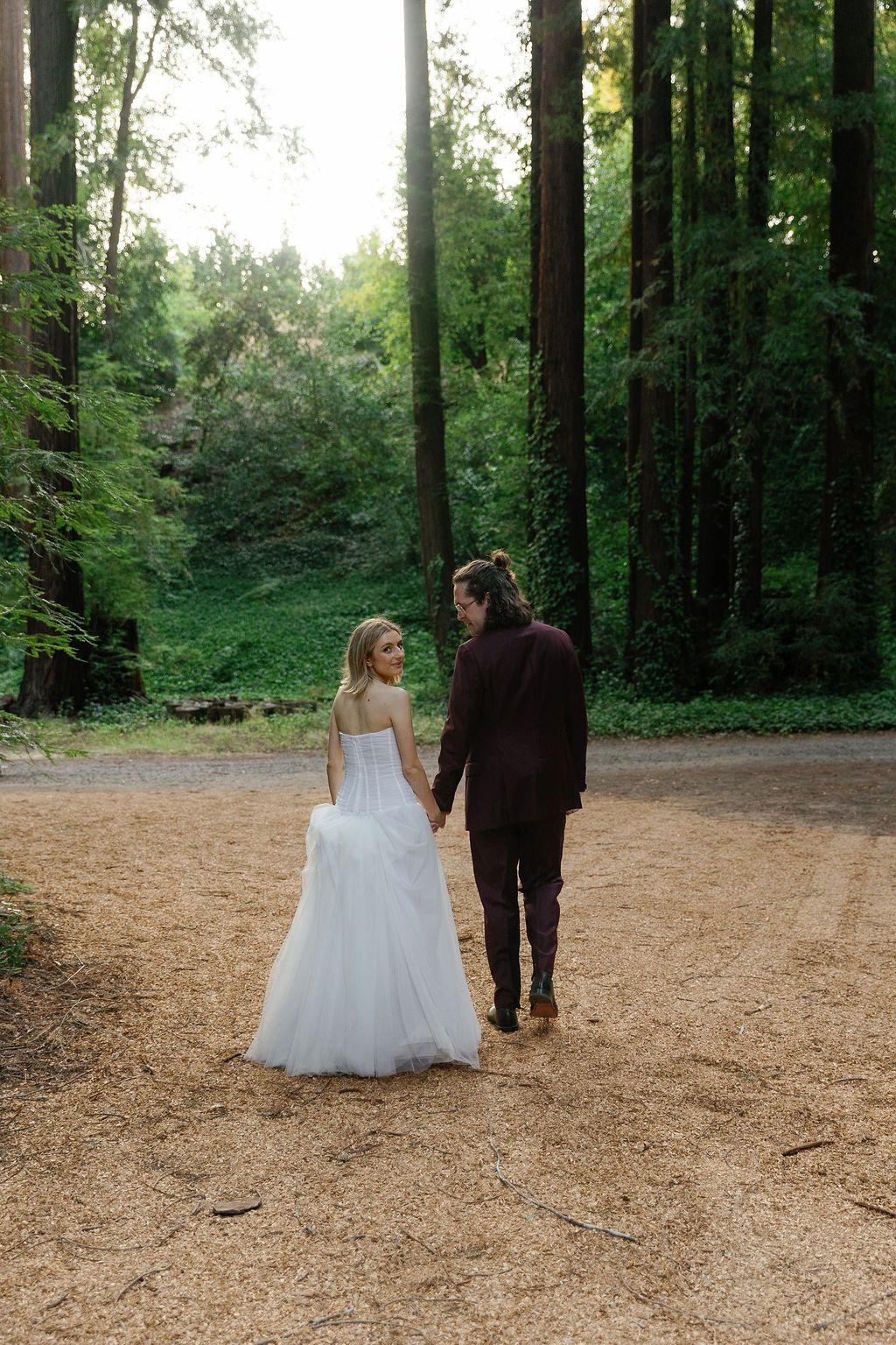 A couple walks through a serene redwood forest clearing on a sunny day.