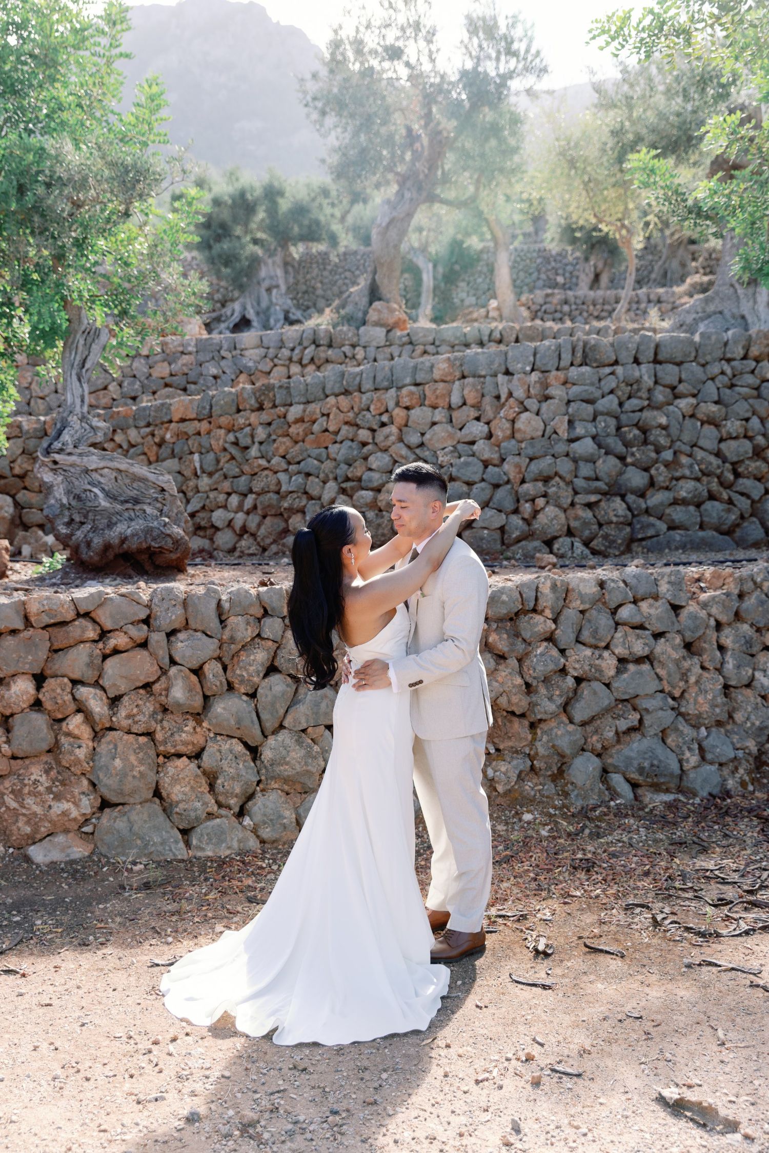 Bride and groom share a romantic dance outdoors near a rustic stone wall surrounded by lush green trees.