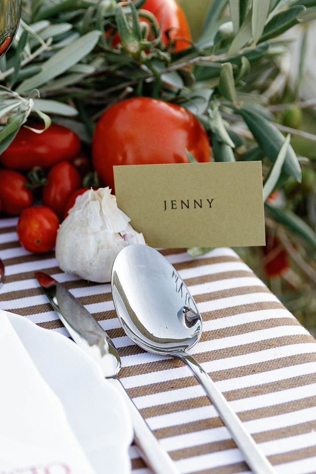 Place card settings on a brown and white striped table runner with garlic bulbs and fresh tomatoes as decor in Tuscany, Italy.