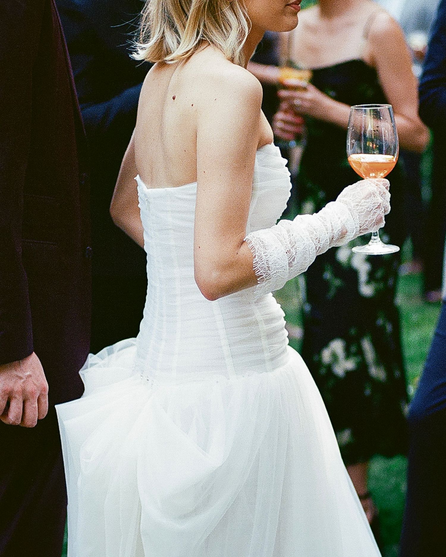 Bride in strapless white gown holding a glass of rosé wine, wearing lace gloves at an outdoor wedding reception.