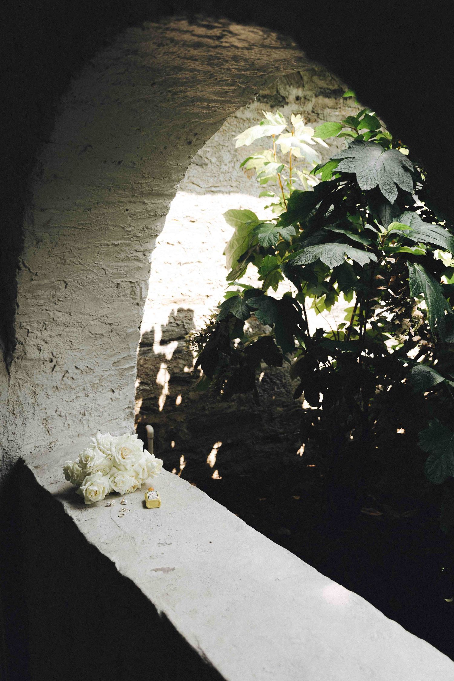 White stone archway leads into a shadowy alcove with green foliage and delicate white flowers creating a romantic garden scene.