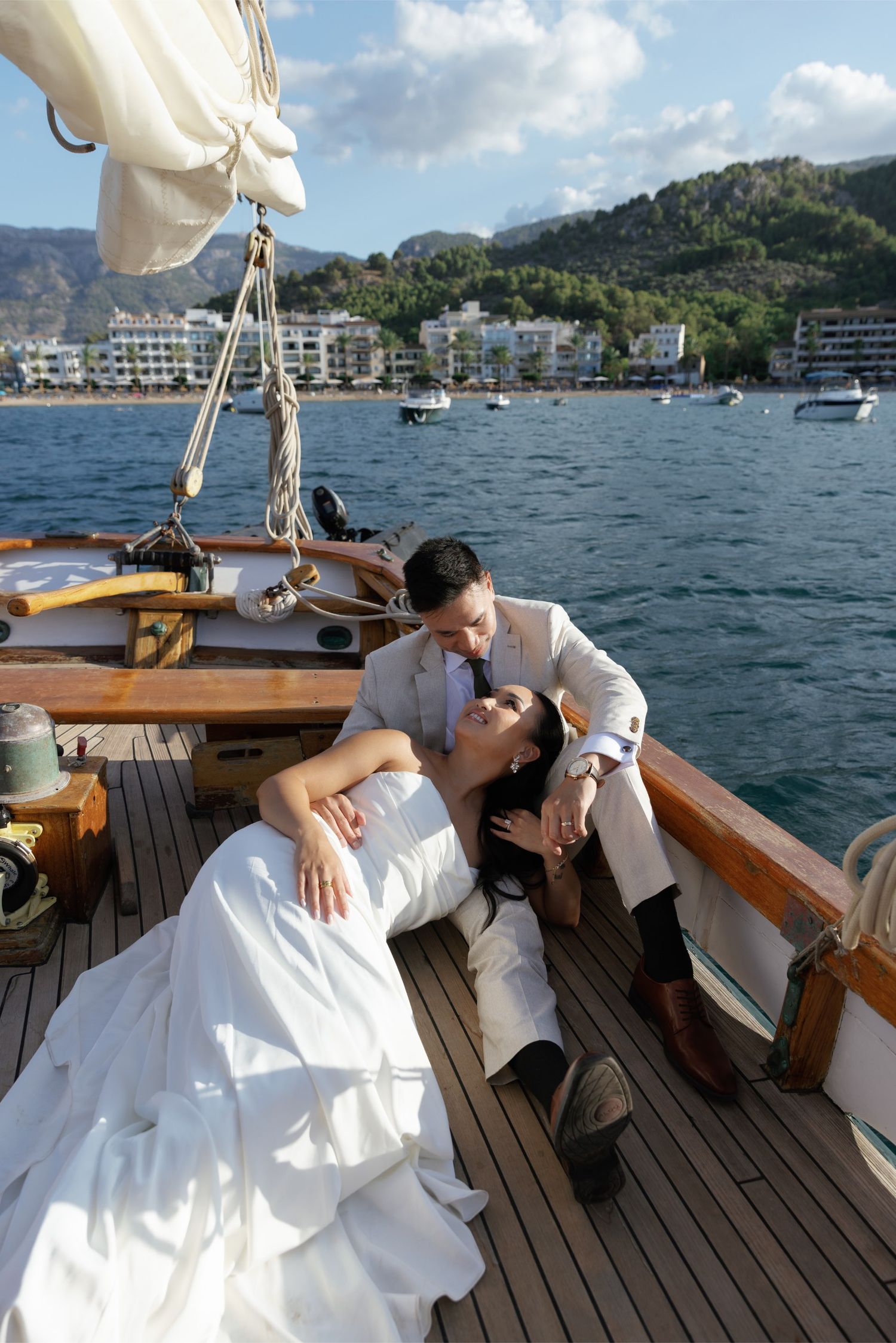 Bride and groom share romantic moment on wooden sailboat deck with Mediterranean coastal town in background.