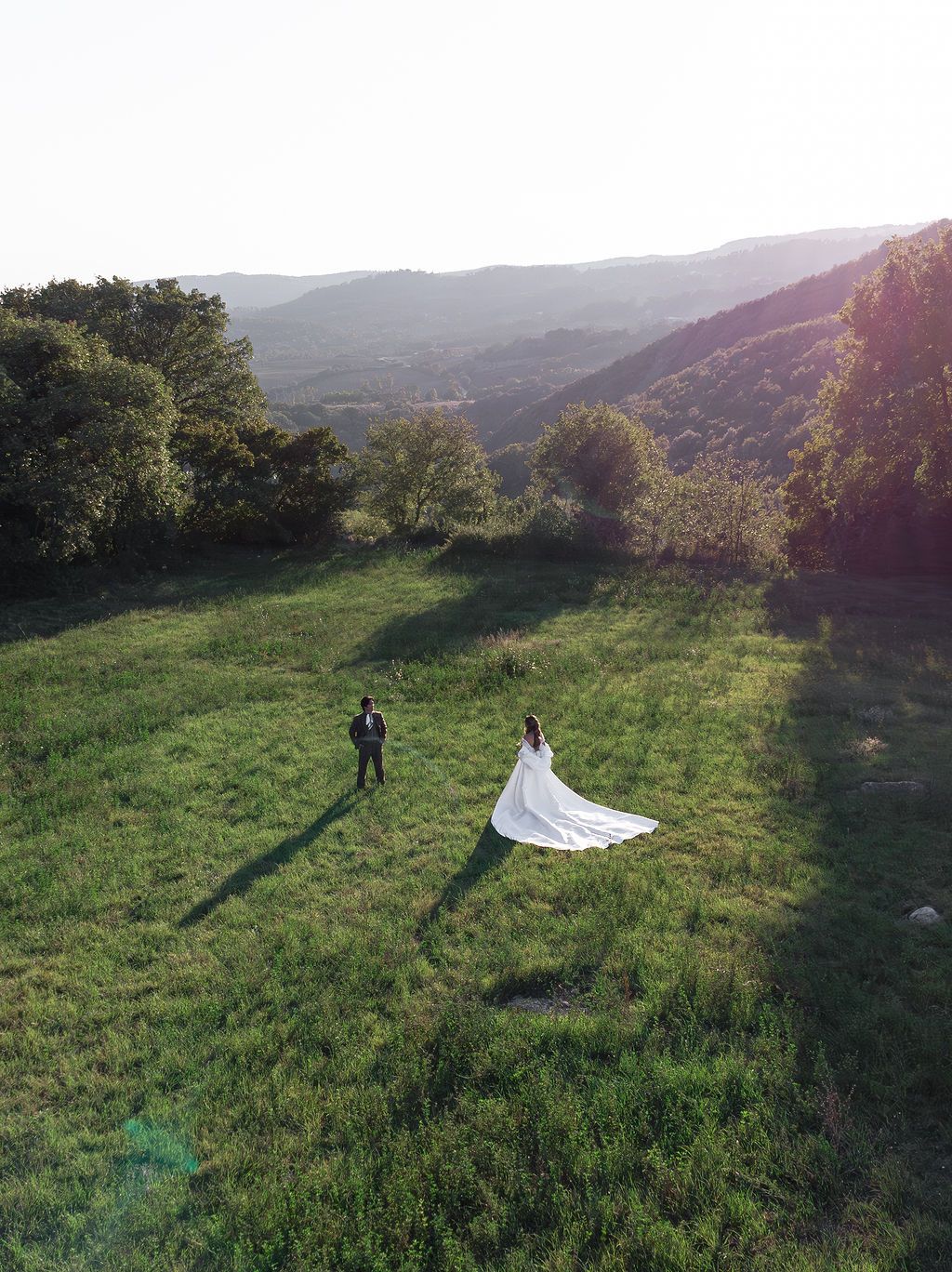 Wedding couple stands in distance on scenic hilltop meadow with mountains in background in Tuscany, Italy.
