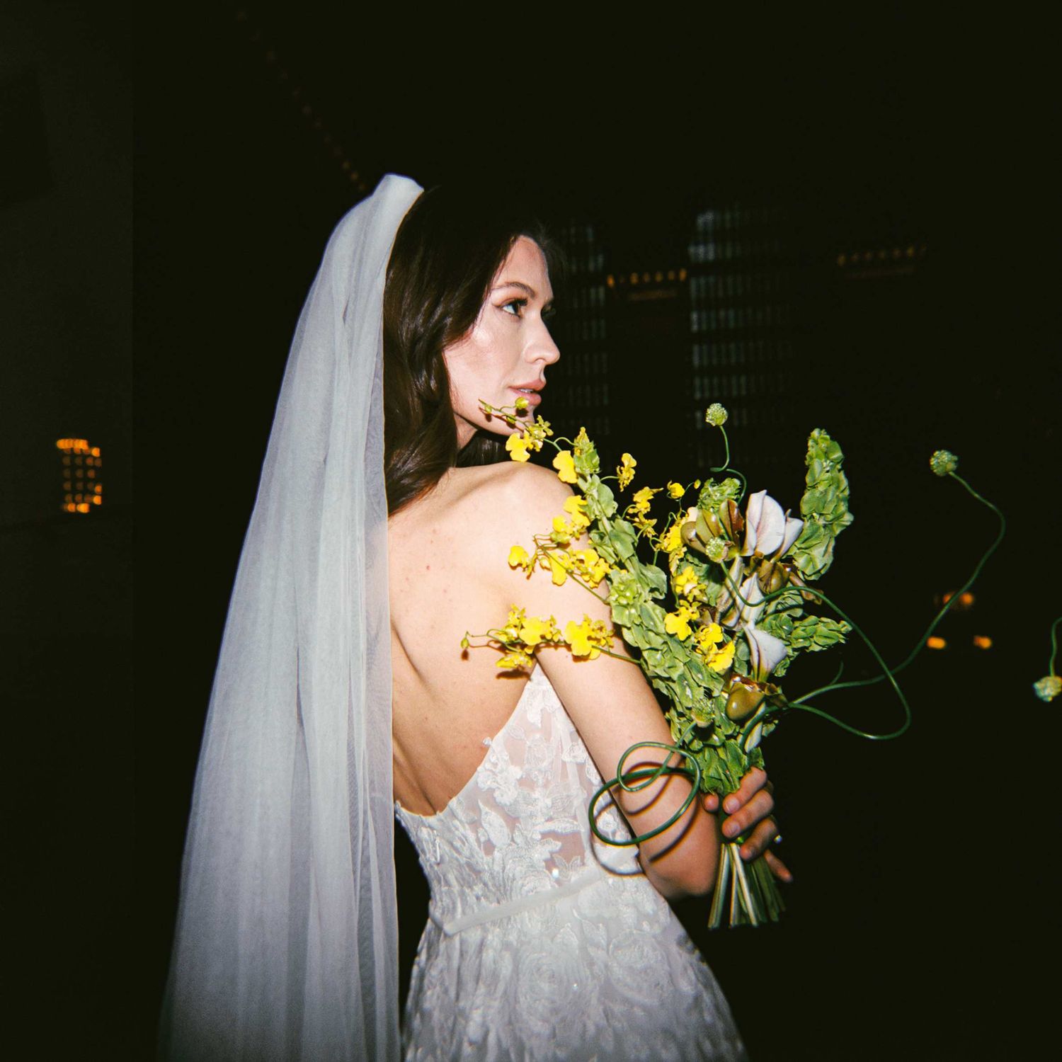 Side view of bride in white lace dress holding yellow flowers, with long white veil against dark background.