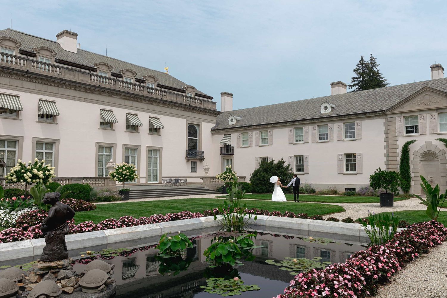 Elegant estate mansion with formal garden, reflecting pool, and wedding couple on lush green grounds.