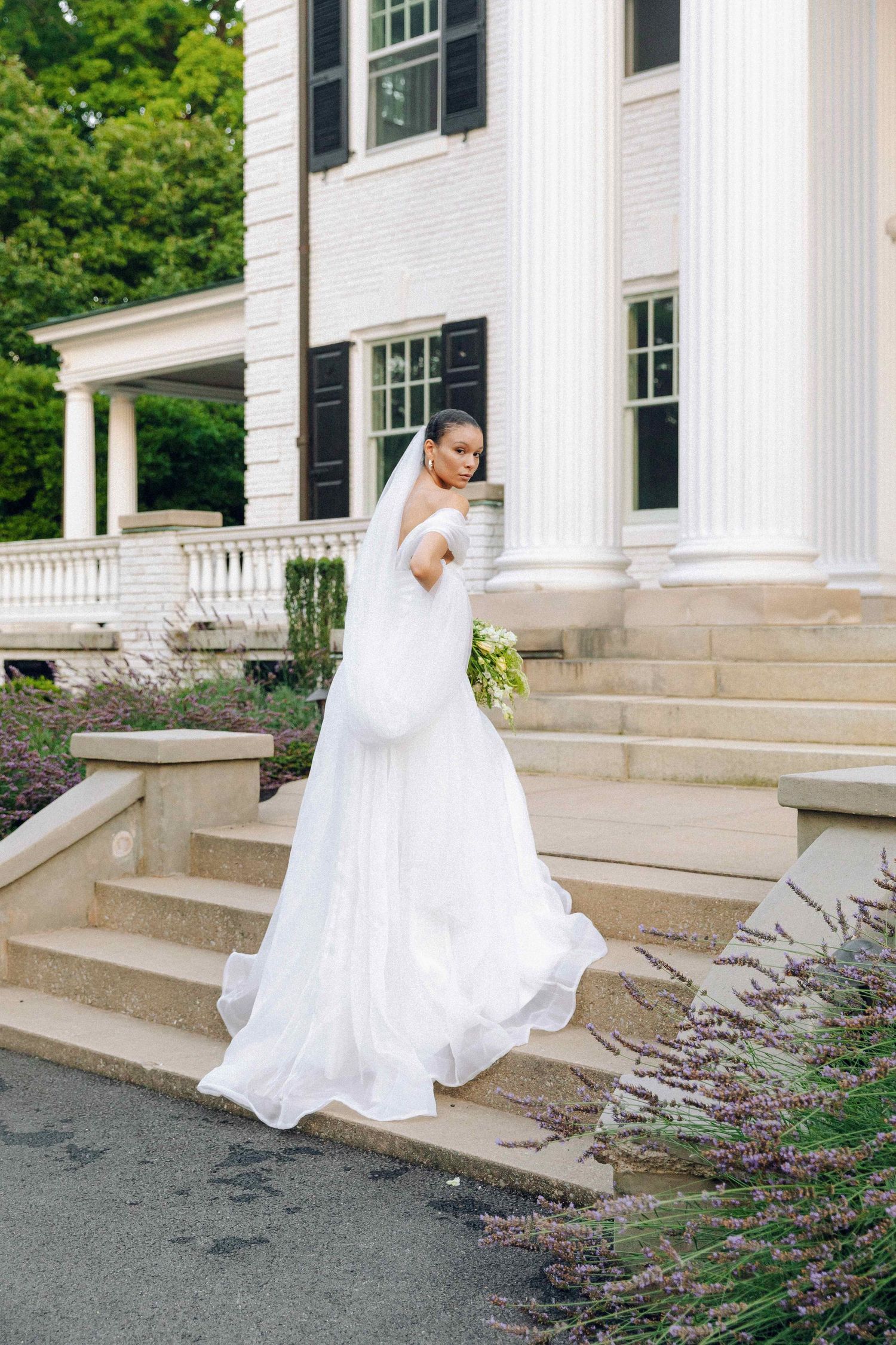 Bride in a flowing white off-shoulder gown with veil holds bouquet on steps of elegant white colonial mansion.