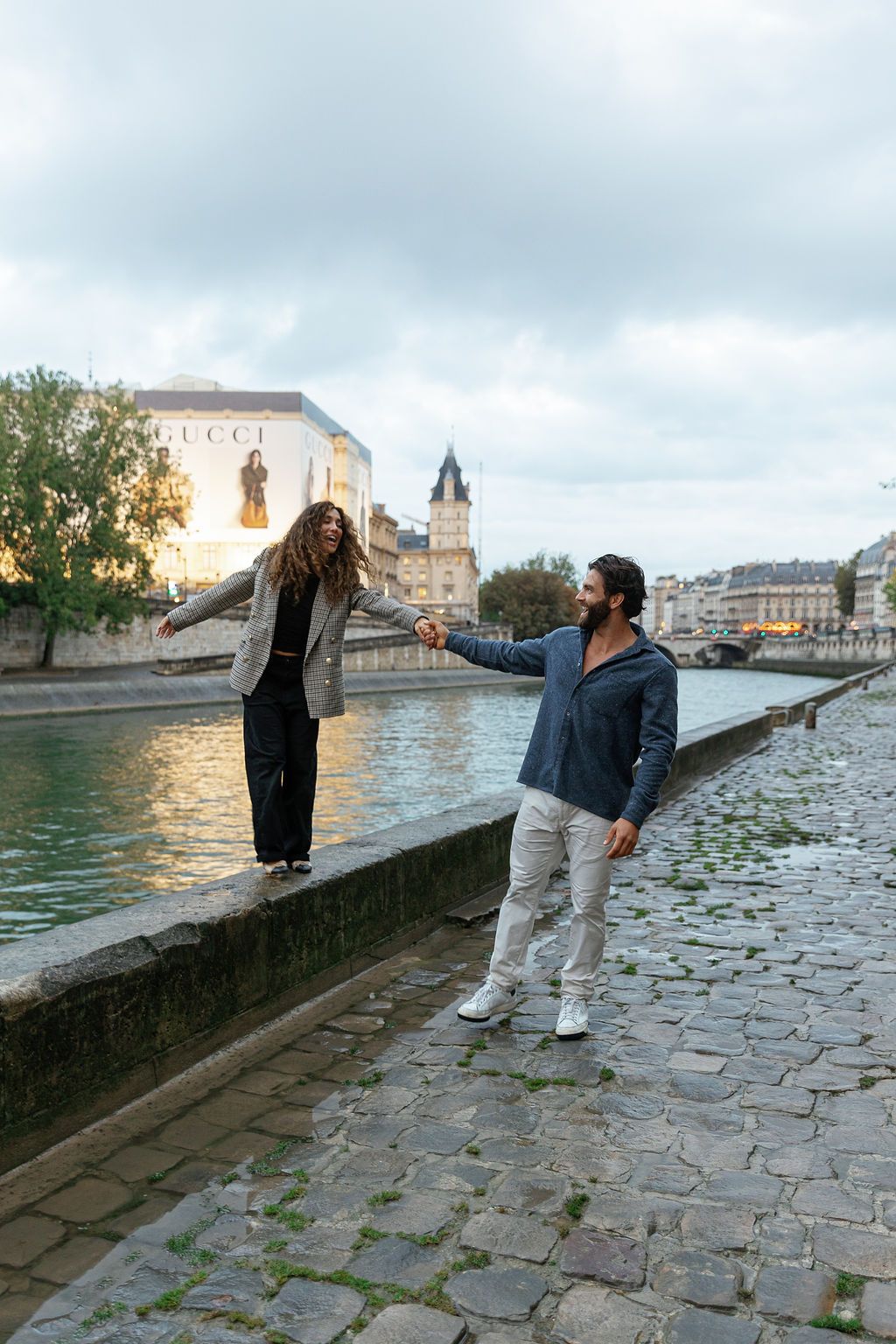 Two people walk along a historic stone riverbank in Paris at sunset as clouds gather overhead.