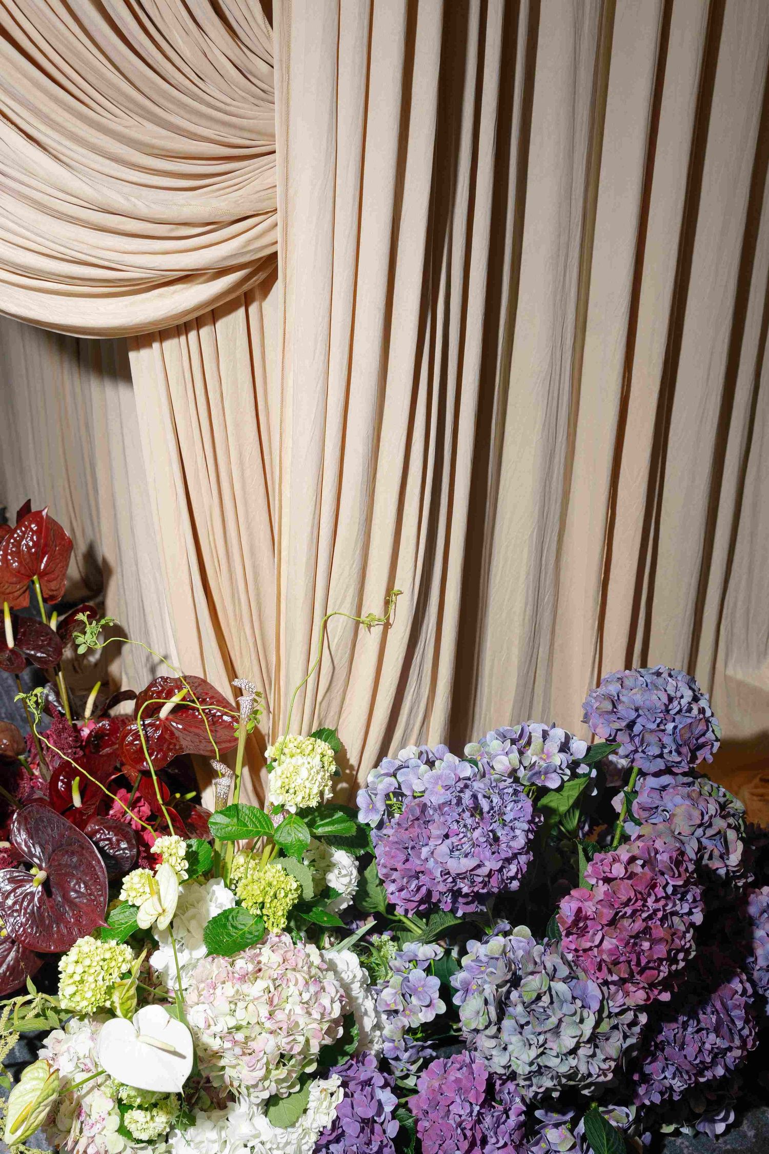 Elegant floral arrangement with purple hydrangeas and mixed flowers against a draped cream backdrop.