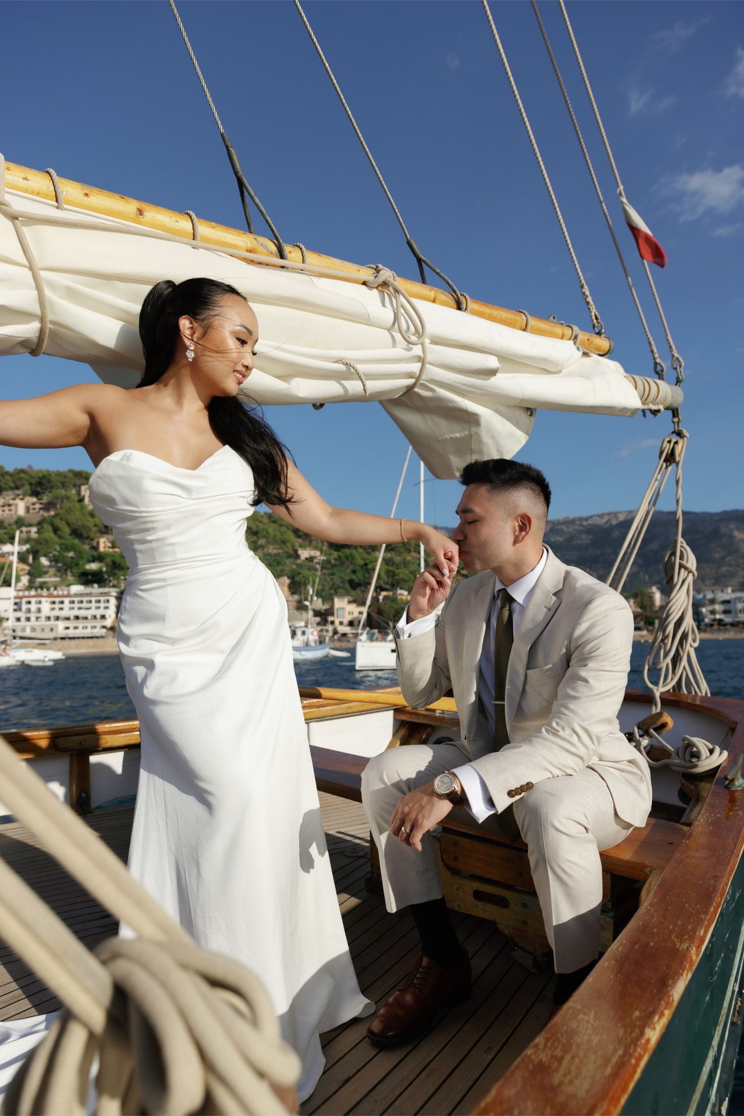 Bride in white strapless gown holds groom's hand on a classic sailboat with Mediterranean coastal town backdrop.