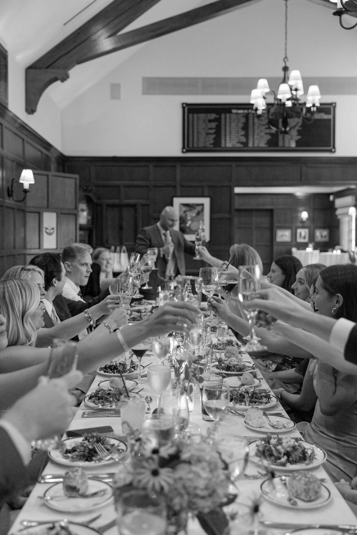 Black and white photo of guests raising glasses in a toast at a long dining table during a formal dinner event.