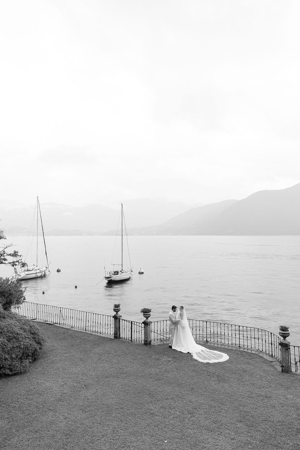 Black and white coastal landscape with boats anchored in a calm harbor viewed from a hillside overlook in Lake Como.