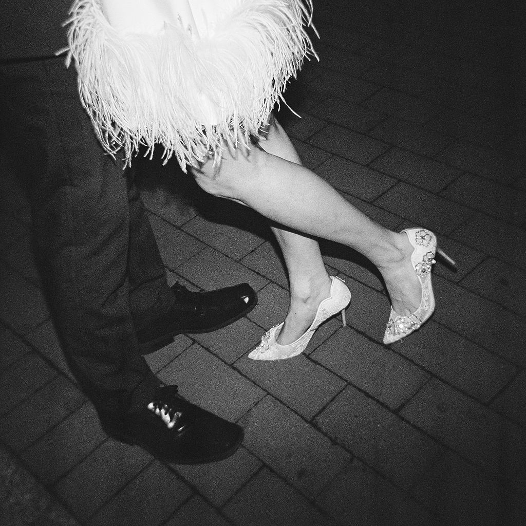 A black and white photo showing legs wearing sparkly heels and a white feathery dress detail on a tiled floor.
