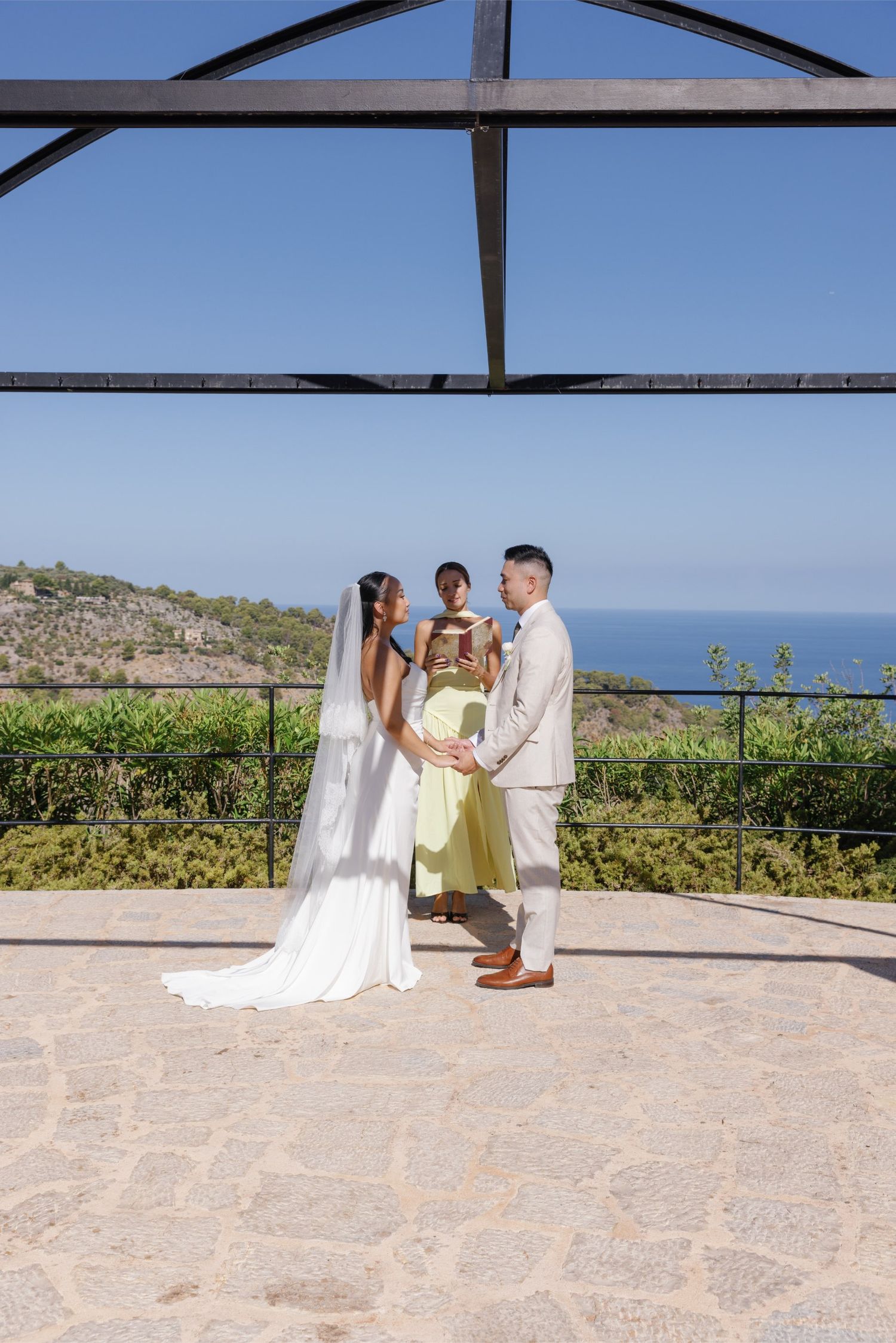 Couple holds hands during outdoor wedding ceremony with ocean view backdrop and officiant in yellow dress.