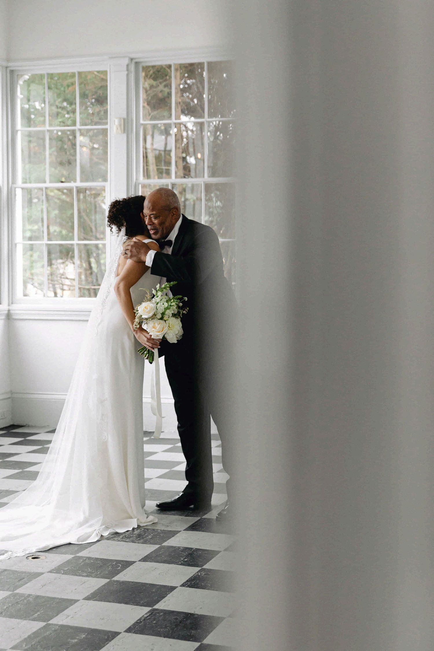 A romantic wedding moment captured in front of bright windows on a black and white checkered floor.