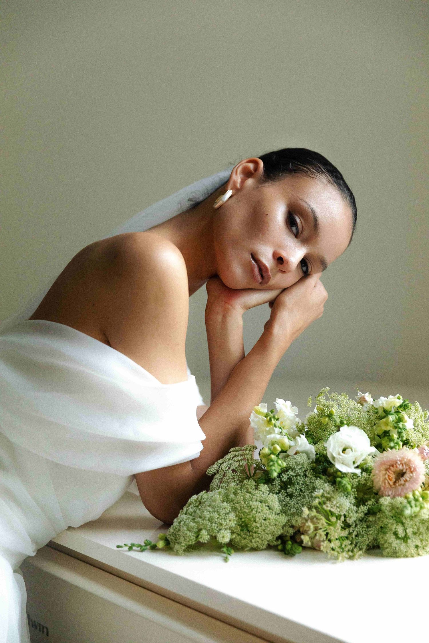 A person in a white drape rests their head near delicate white and green floral arrangements on a light surface.