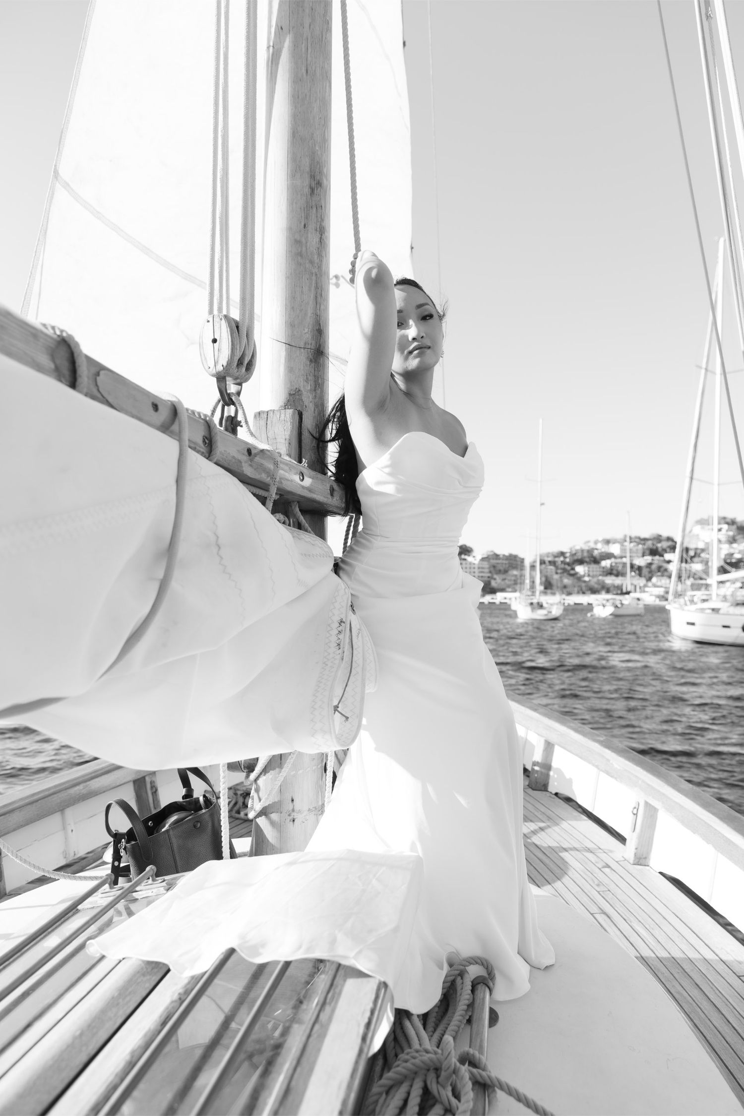 Black and white photo of a bride in a white gown posing on a sailboat deck with sails and ocean in the background.