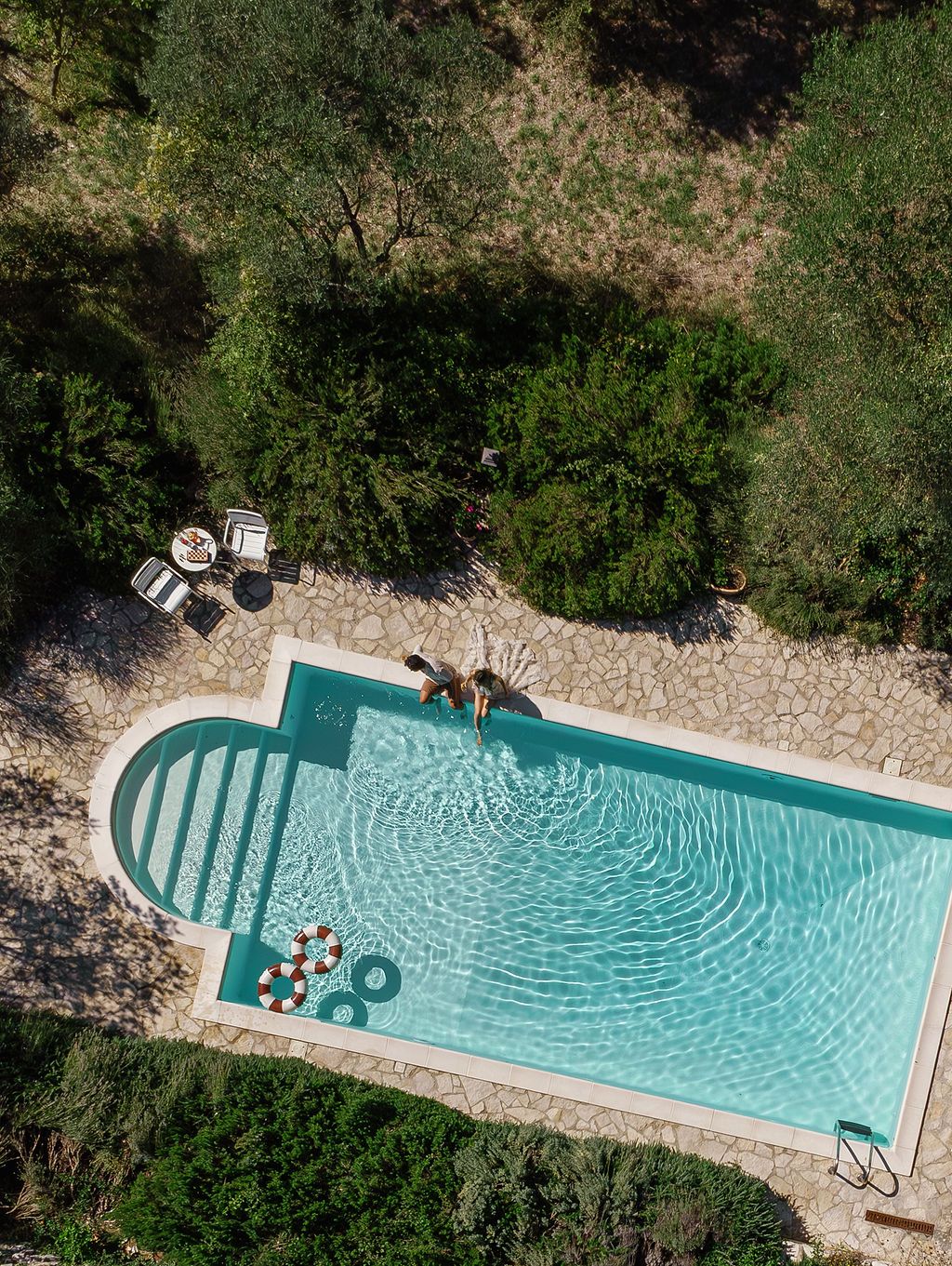 Aerial view of a long turquoise swimming pool surrounded by trees and landscaping with lounge chairs at one end in Tuscany, Italy.