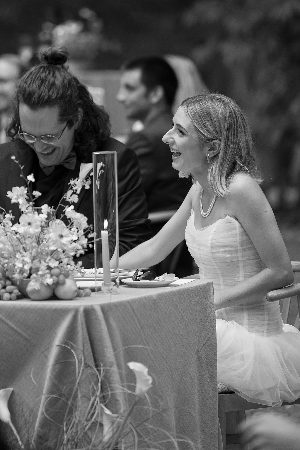Wedding party seated at a long table with white linens and floral centerpieces during an outdoor reception.