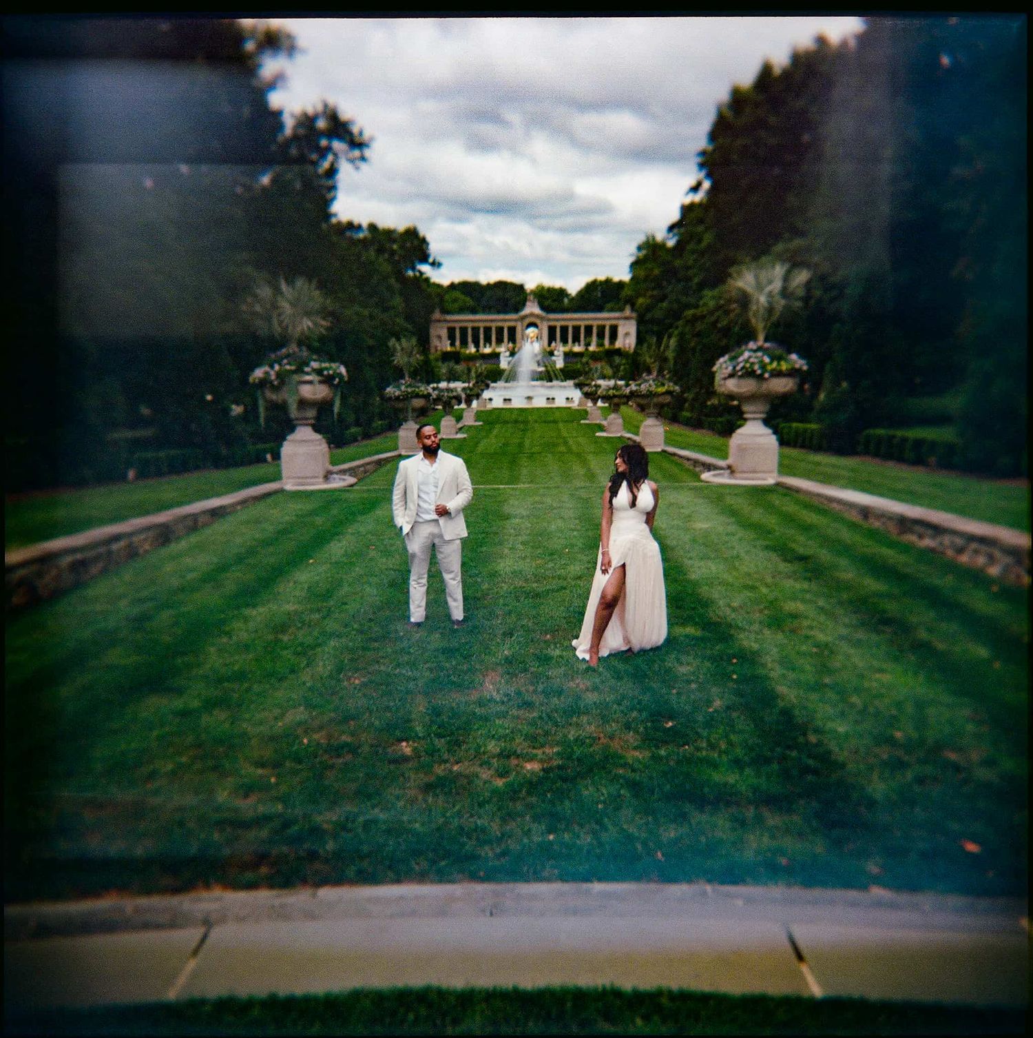 Couple in white attire standing on lush green garden lawn with fountain and pergola backdrop under cloudy sky.
