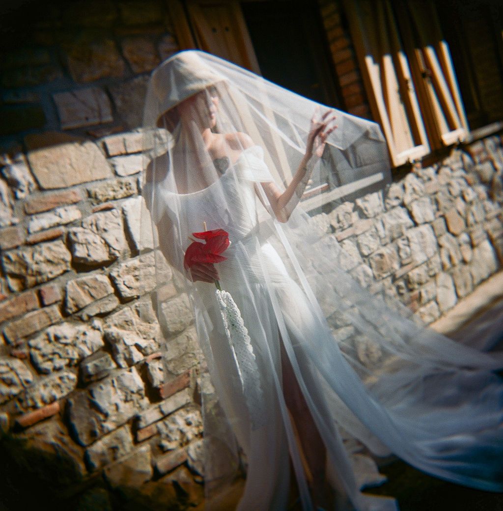 A bride in a flowing white veil and dress holds a red bouquet against a rustic stone wall background in Tuscany, Italy.