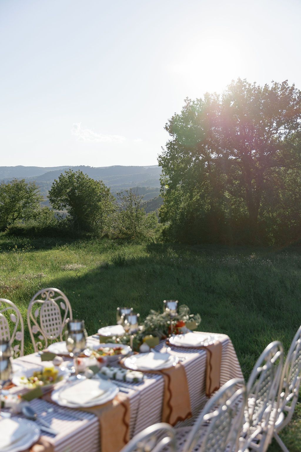 A rustic outdoor dining setup with white chairs and table settings against rolling hills  in Tuscany, Italy.
