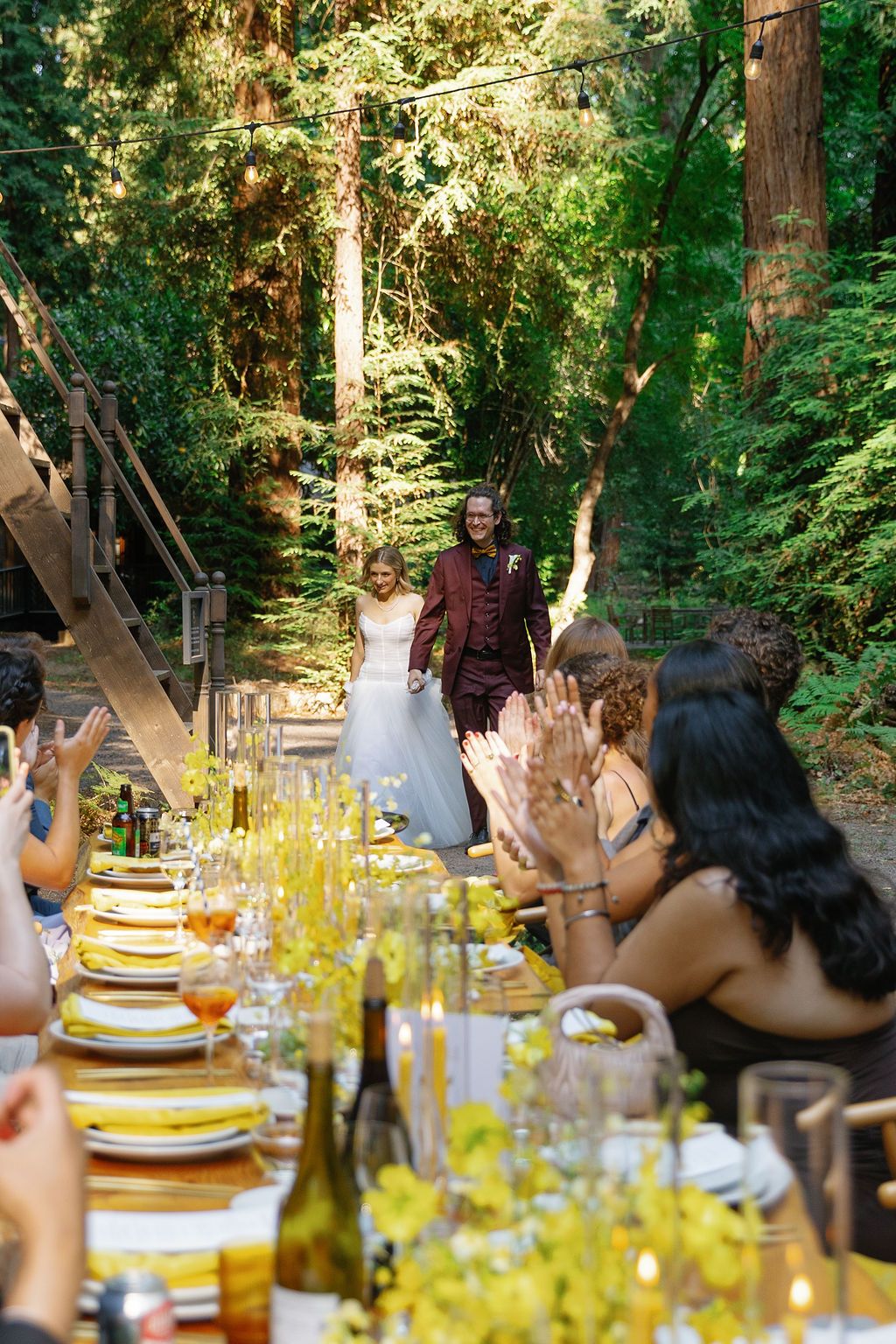 Outdoor dining table set with yellow decor and place settings in a sunlit forest setting.