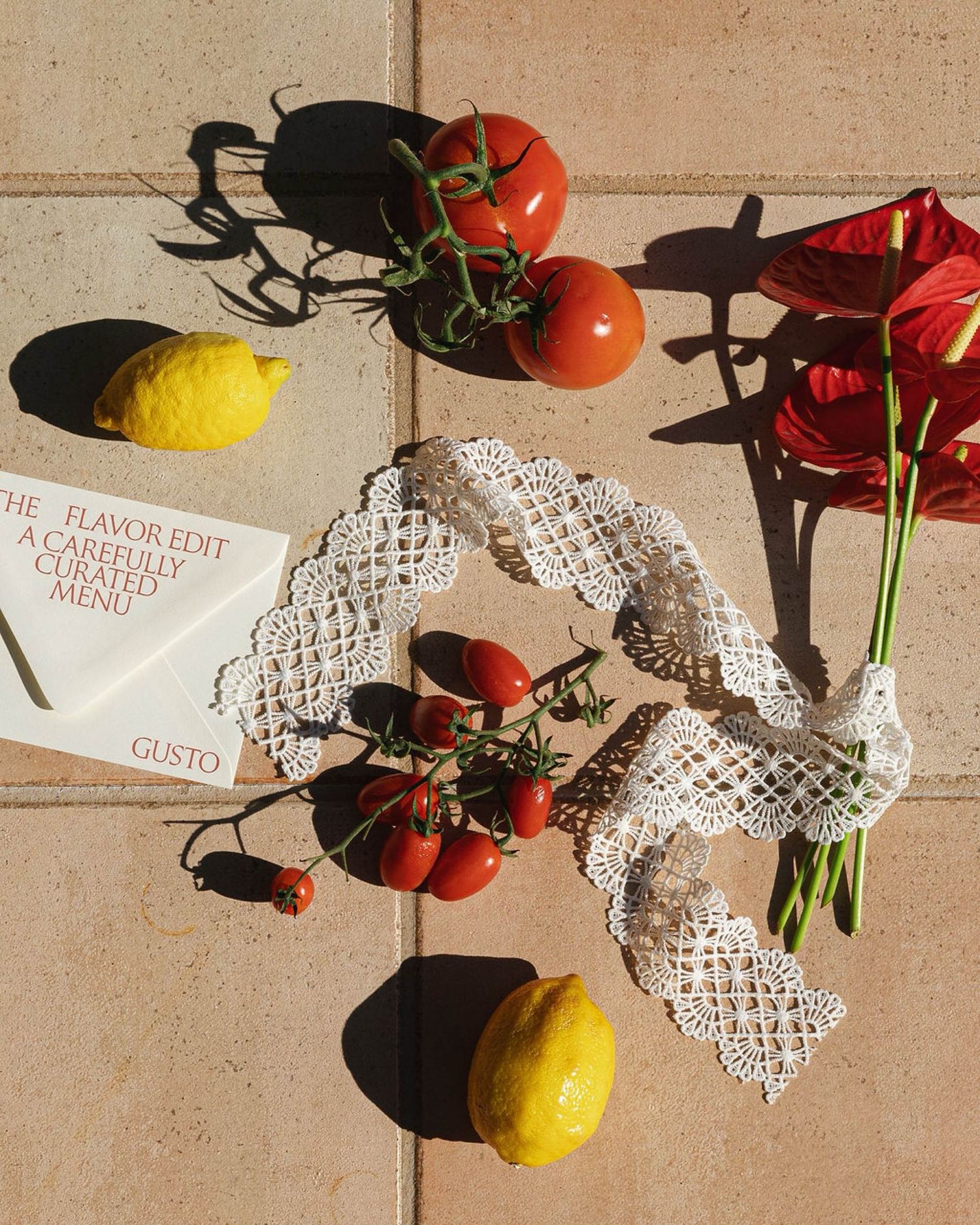 Fresh tomatoes on the vine, lemons, and red chili peppers arranged with lace doily and book on beige tile surface.
