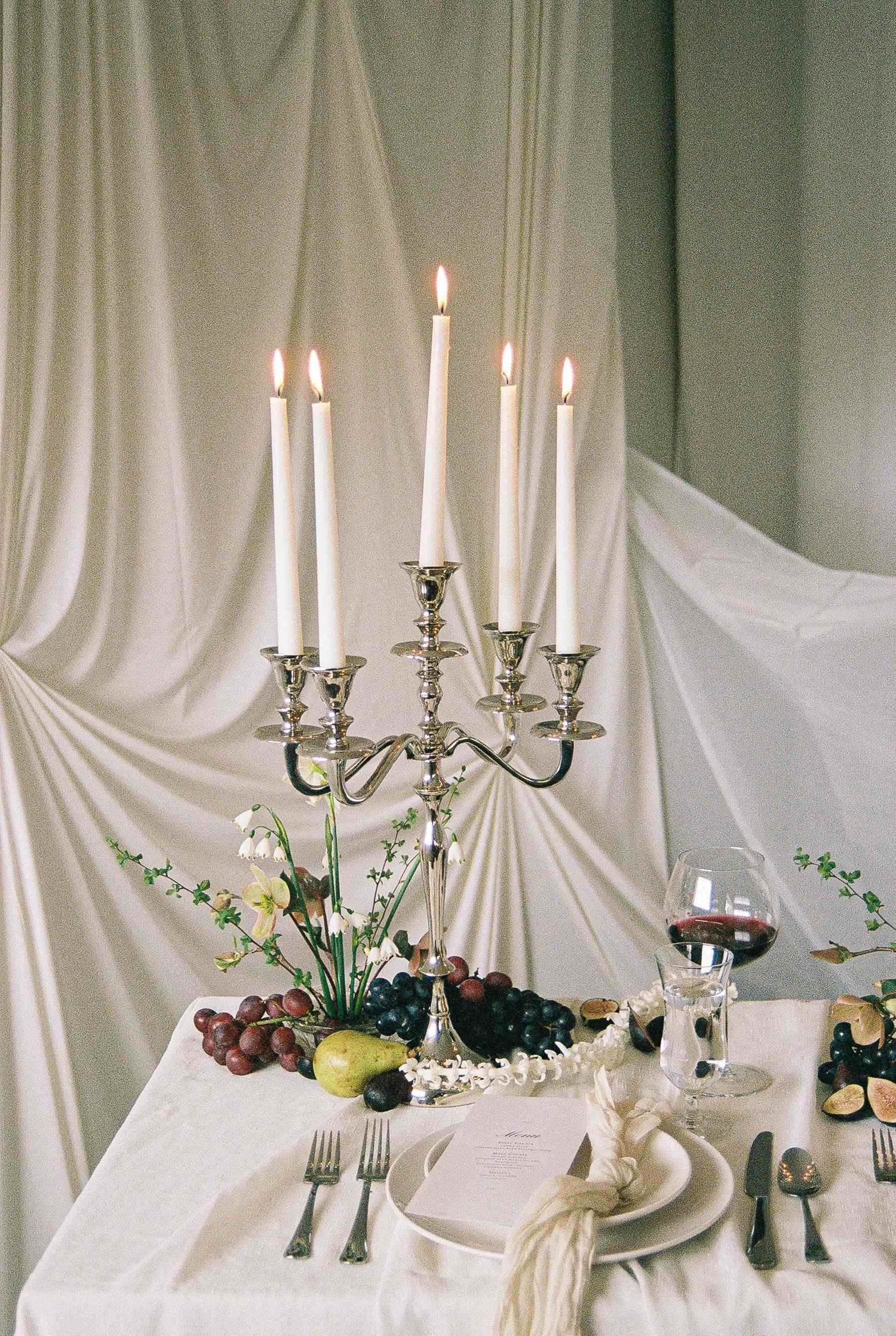 Elegant table setting with silver candelabra, wine glass, and fruit arrangement against draped white fabric backdrop.