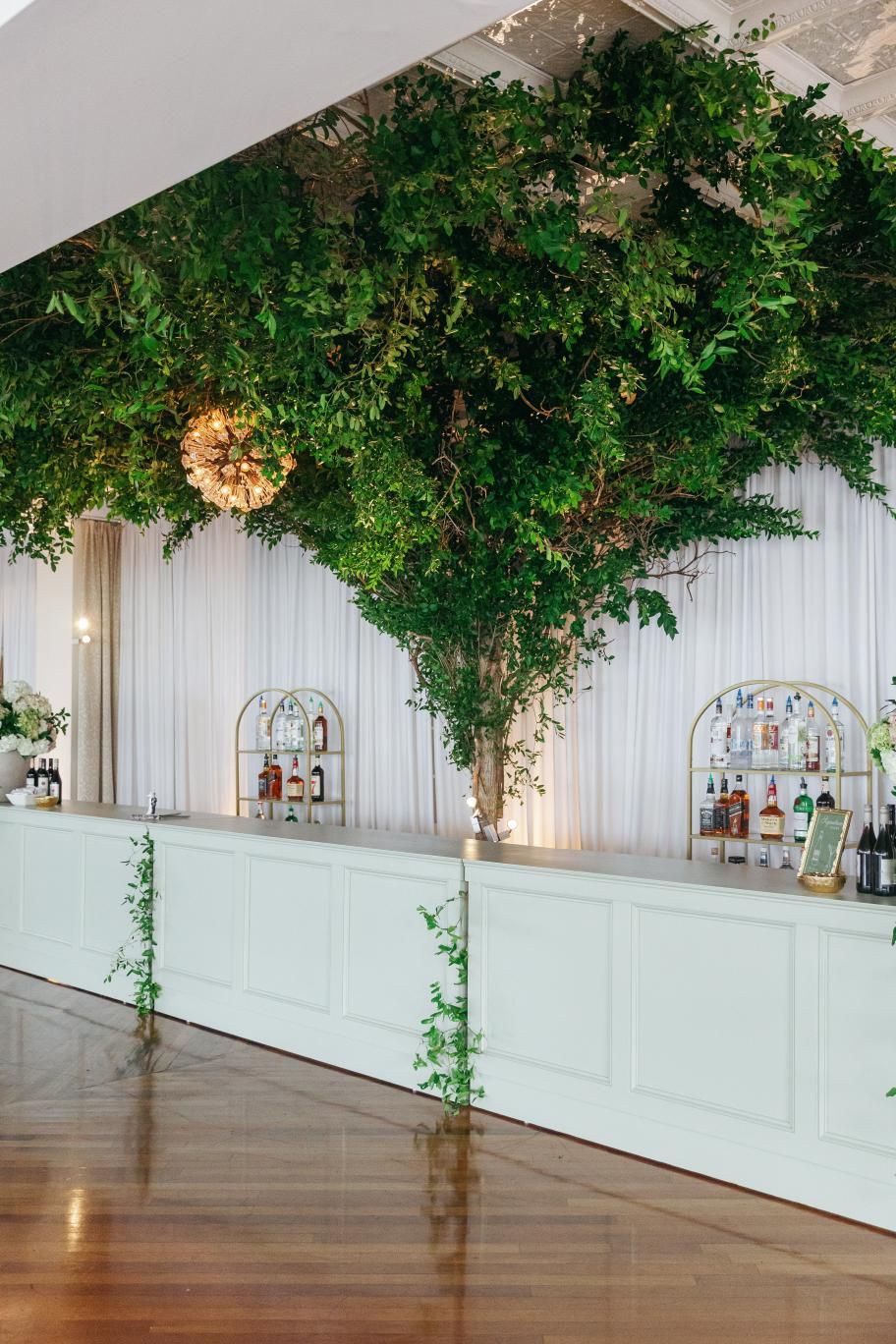 A series of venue photos showing large green floral installations hanging from the ceiling with white wainscoting walls.