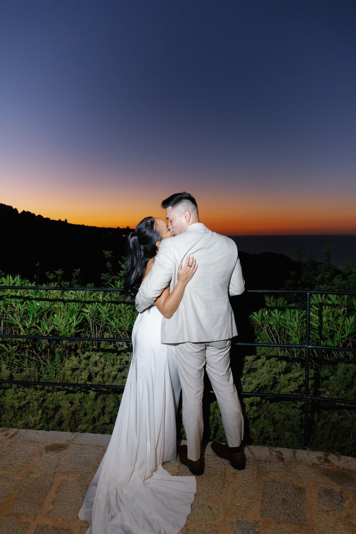 Newlyweds share romantic kiss at sunset overlooking lush landscape, bride in white gown, groom in beige suit.