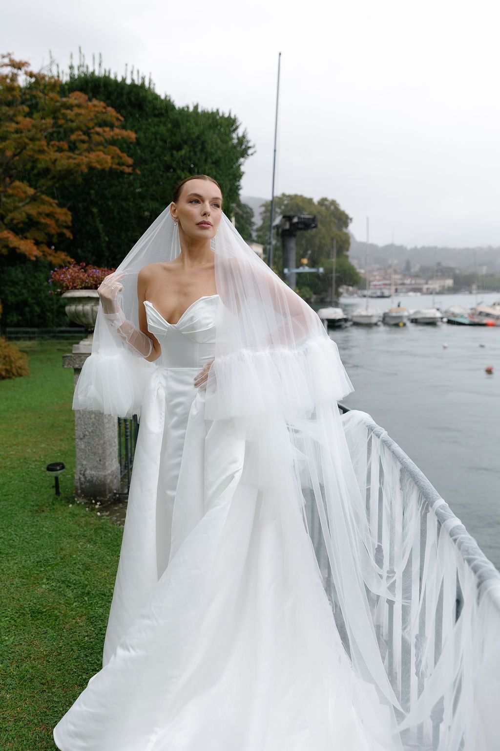 A bride in an elegant white wedding gown and flowing veil stands by a waterfront railing on an overcast day in Lake Como.