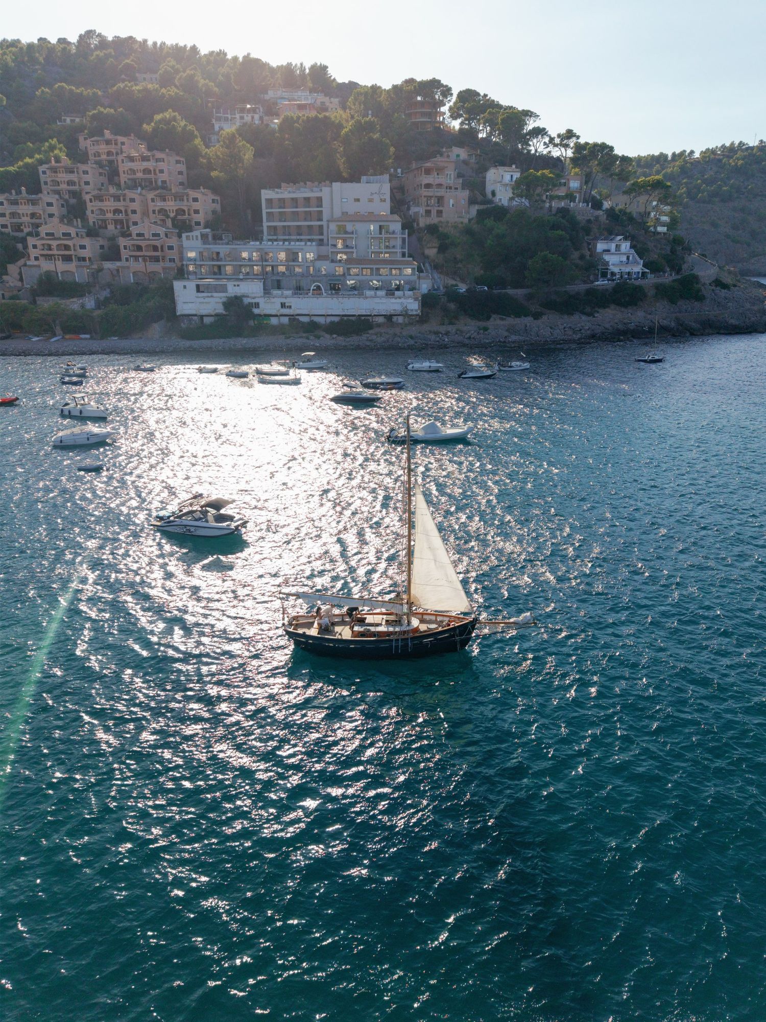 Aerial view of a sailboat on turquoise Mediterranean waters near a hillside coastal village on a sunny day.