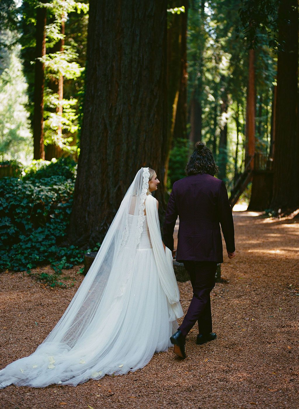 A flowing white wedding dress train trails behind on a forest path lined with towering redwoods.