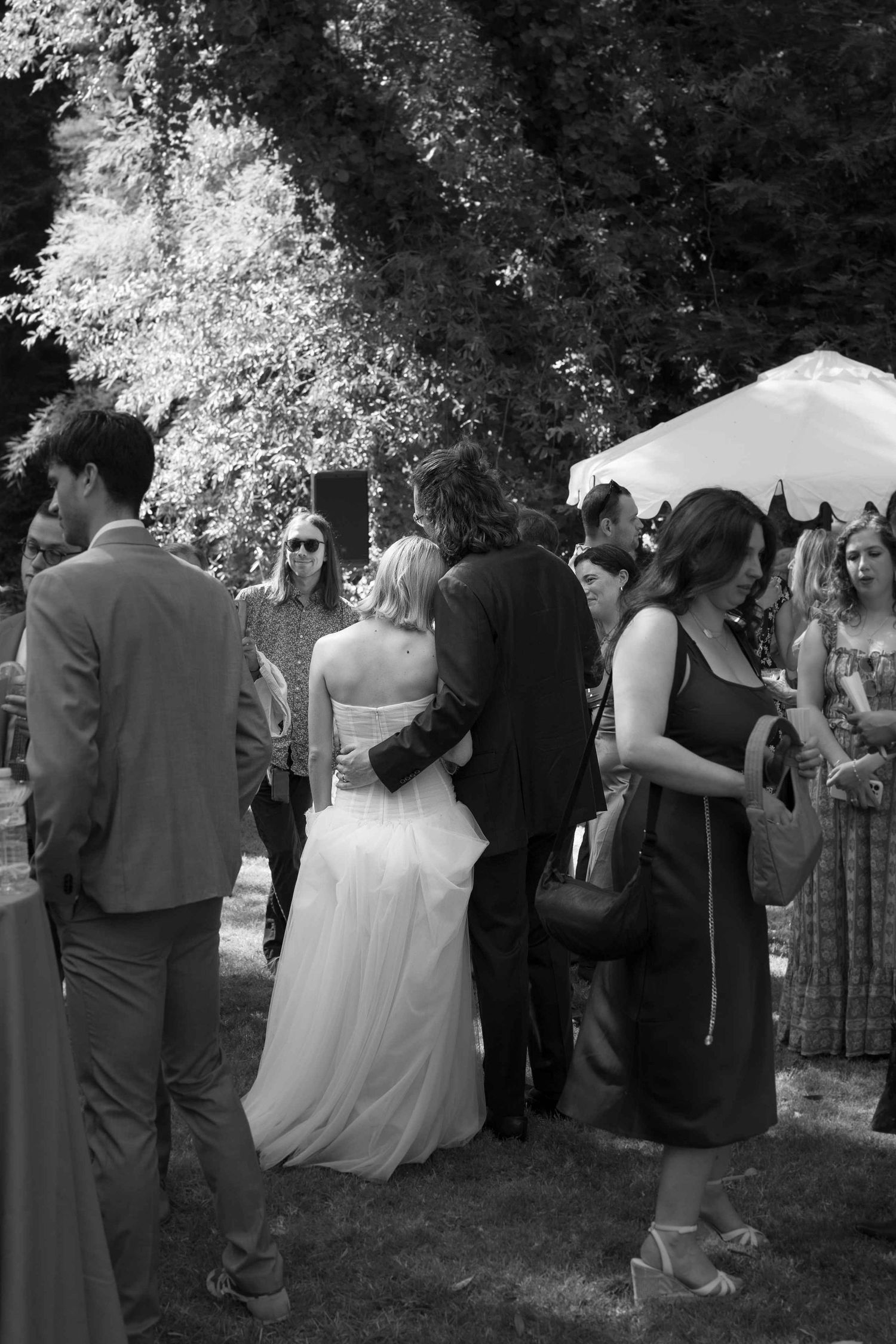 Black and white outdoor wedding reception with bride in white gown embraced by groom among elegantly dressed guests.