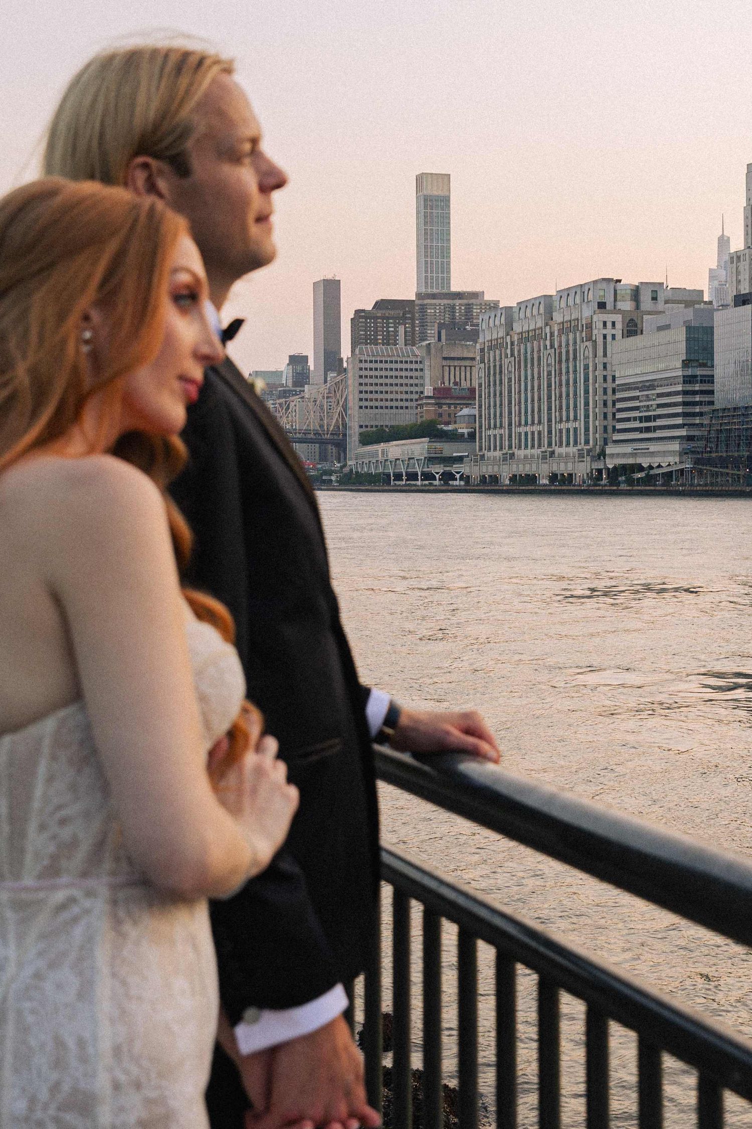 A couple looks out over the Manhattan skyline and East River from a waterfront railing at dusk.