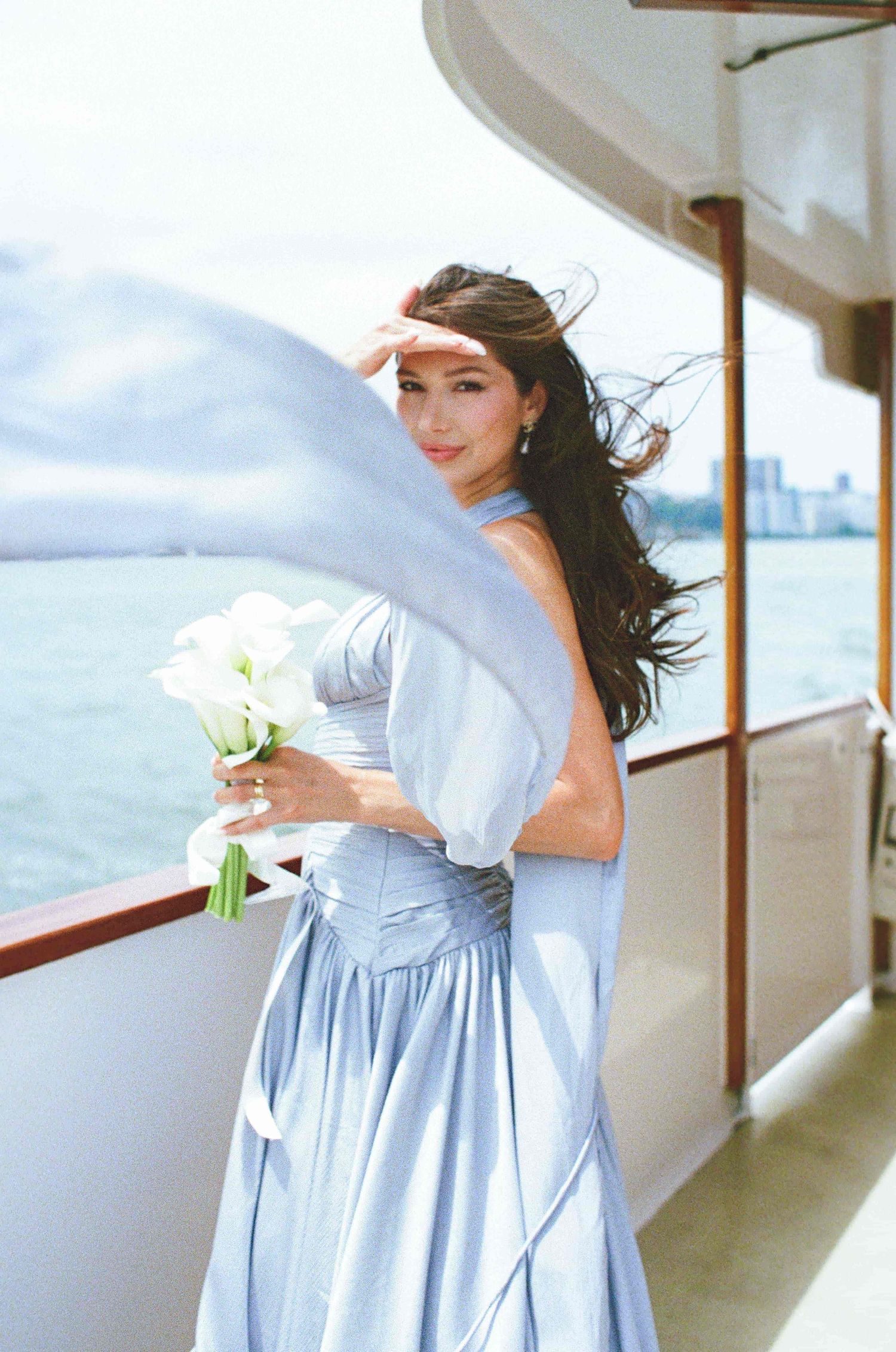 Woman in blue gown holding white calla lily bouquet on yacht deck, hair blowing in wind with city skyline backdrop.