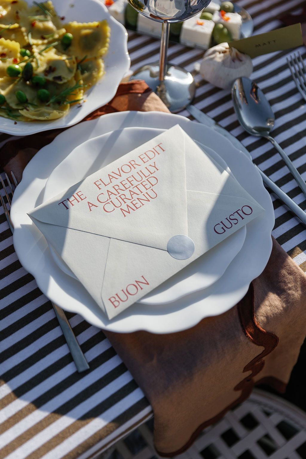 White place setting with menu card reading 'Buon Gusto' on a striped tablecloth in sunlight in Tuscany, Italy.