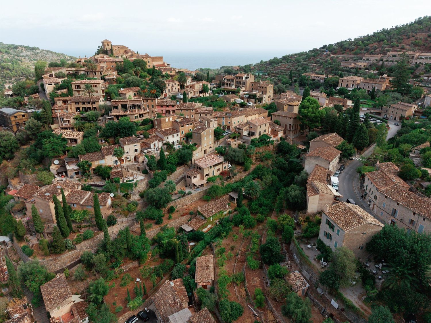 Aerial view of a historic Mediterranean hillside village with stone buildings surrounded by lush greenery.