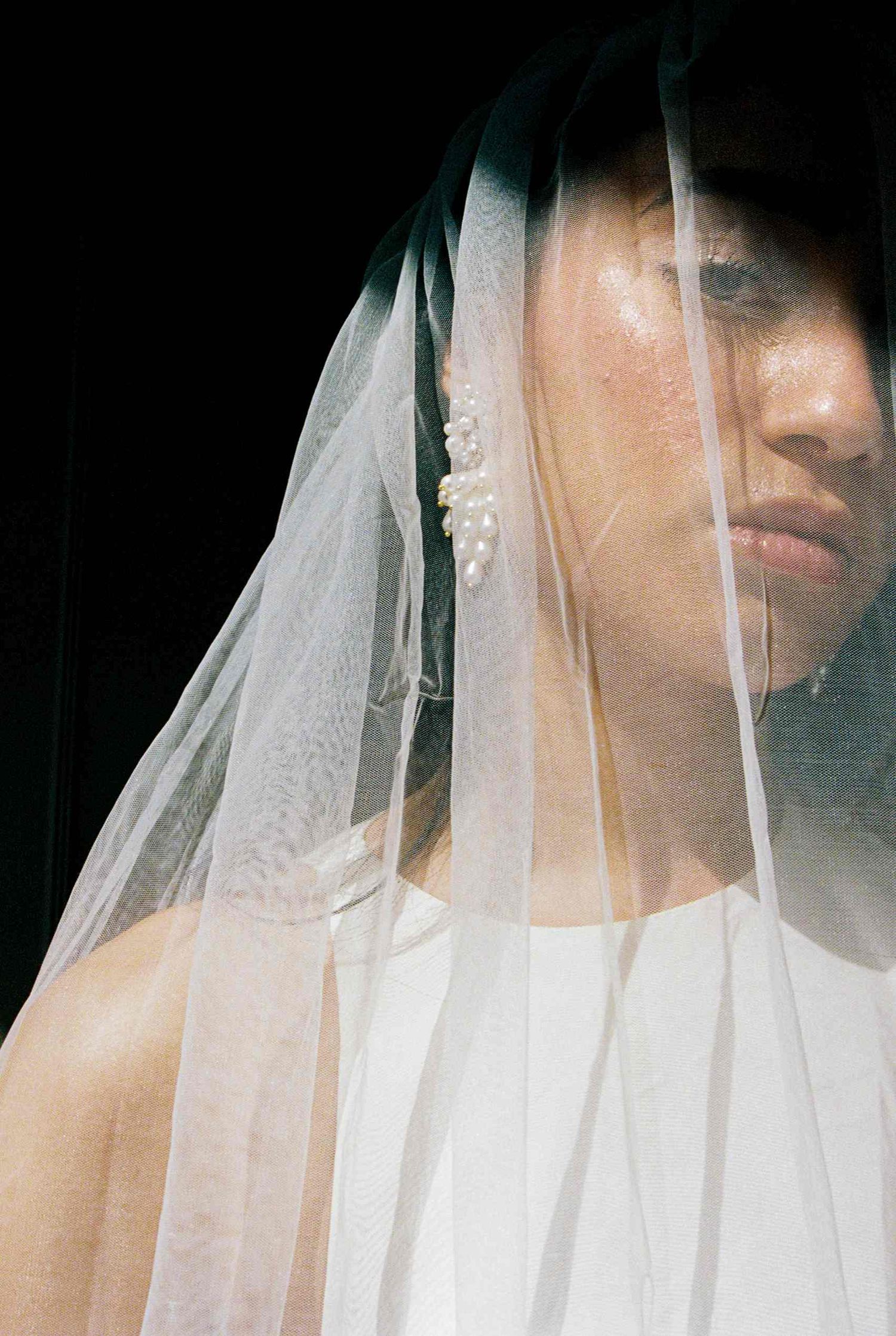 A delicate white bridal veil drapes over someone wearing pearl earrings in a moody, artistic portrait.