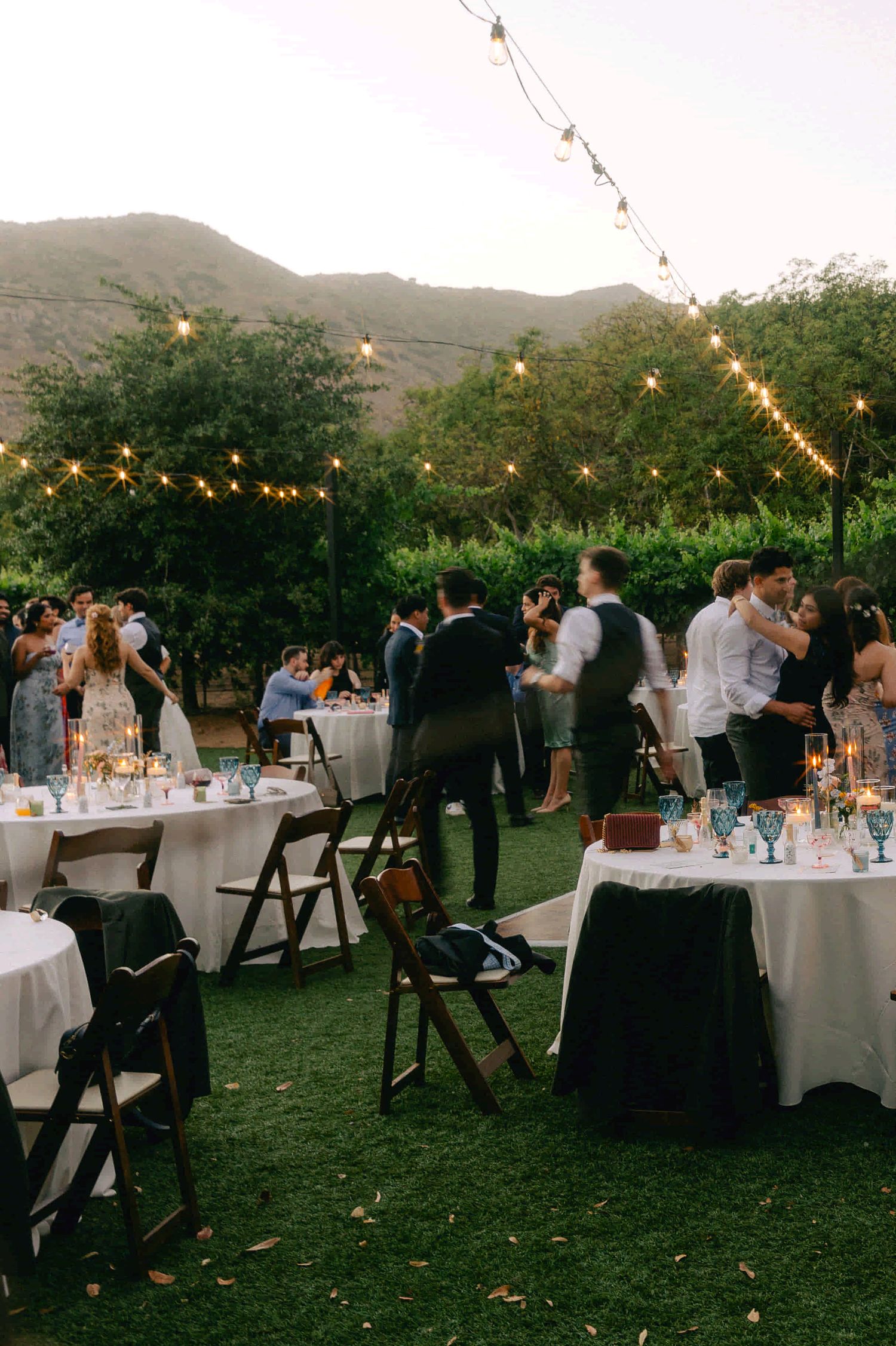 Outdoor wedding reception at sunset with string lights illuminating round tables on a grassy lawn near mountains.