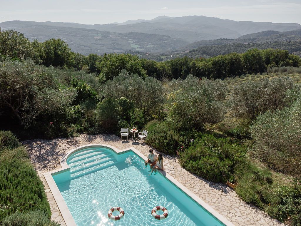 Aerial views of a turquoise swimming pool surrounded by lush greenery and rolling hills in the countryside in Tuscany, Italy.