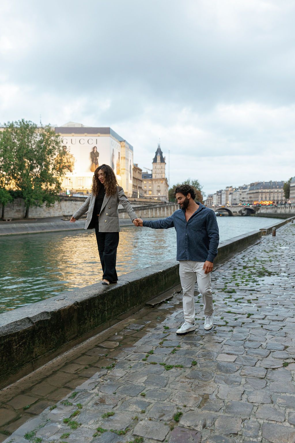 Two people walking along the Seine River in Paris with Notre Dame cathedral visible in the background at sunset.