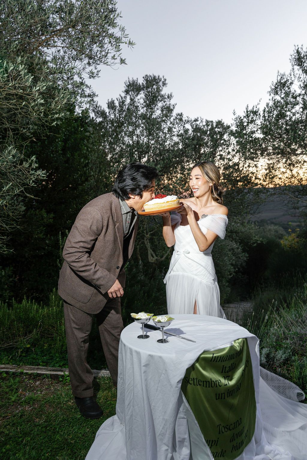 A couple in wedding attire shares a playful moment drinking from bottles in an outdoor garden setting at sunset.