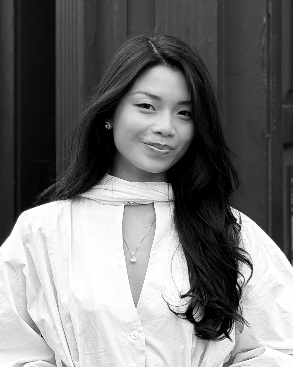 Black and white portrait of a young woman with long wavy hair wearing a white blouse and delicate necklace.