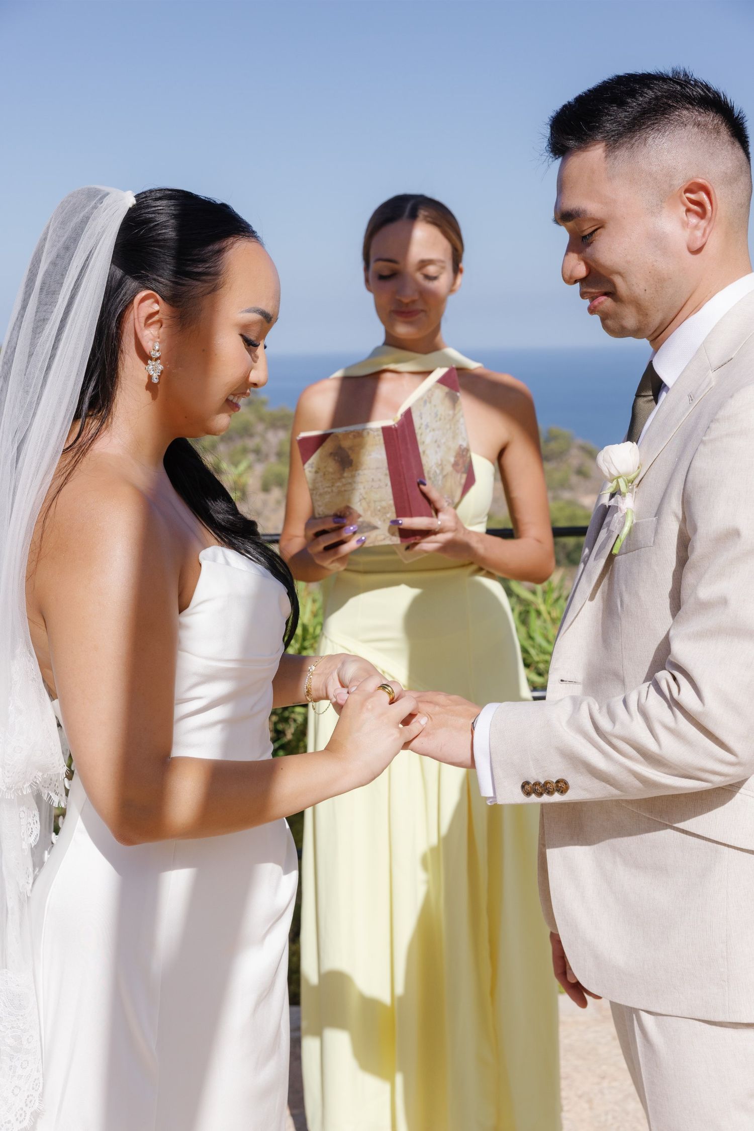Bride and groom exchange rings during outdoor wedding ceremony with ocean view, officiant holding book in background.