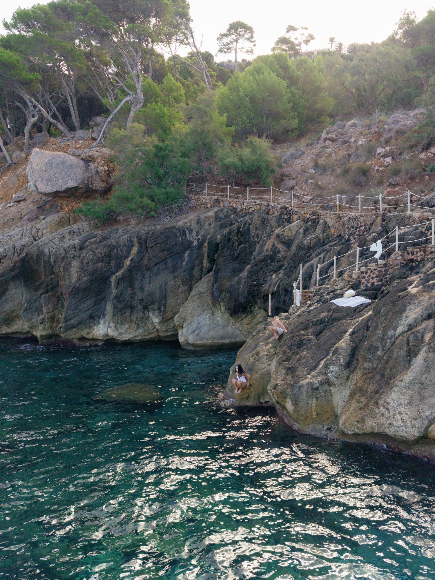 Rocky coastal cliffs with turquoise water, wooden railings on a narrow path, and lush green trees overlooking a secluded cove.