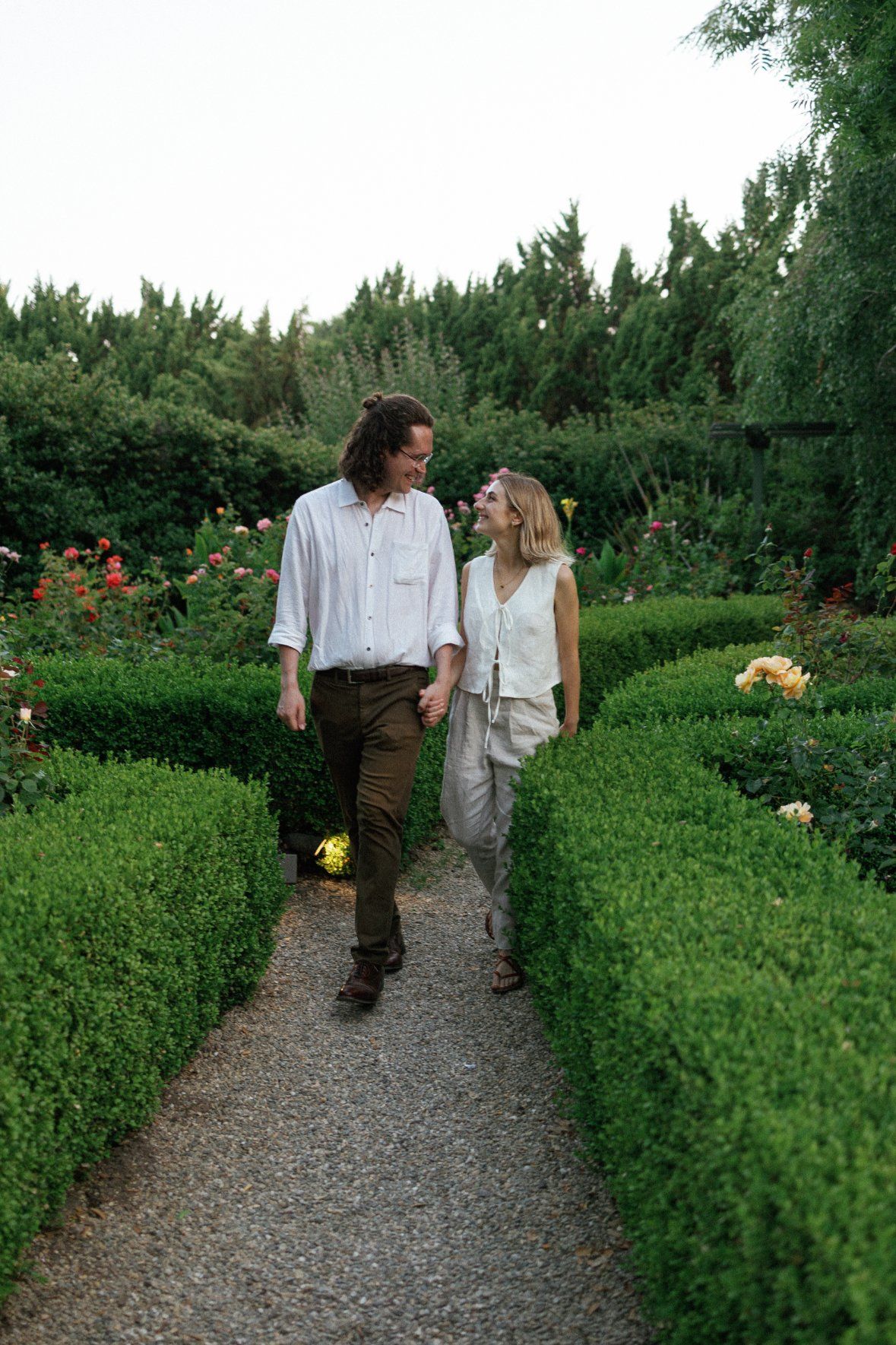 A couple walks together on a gravel path between neatly trimmed hedges in a lush garden setting.