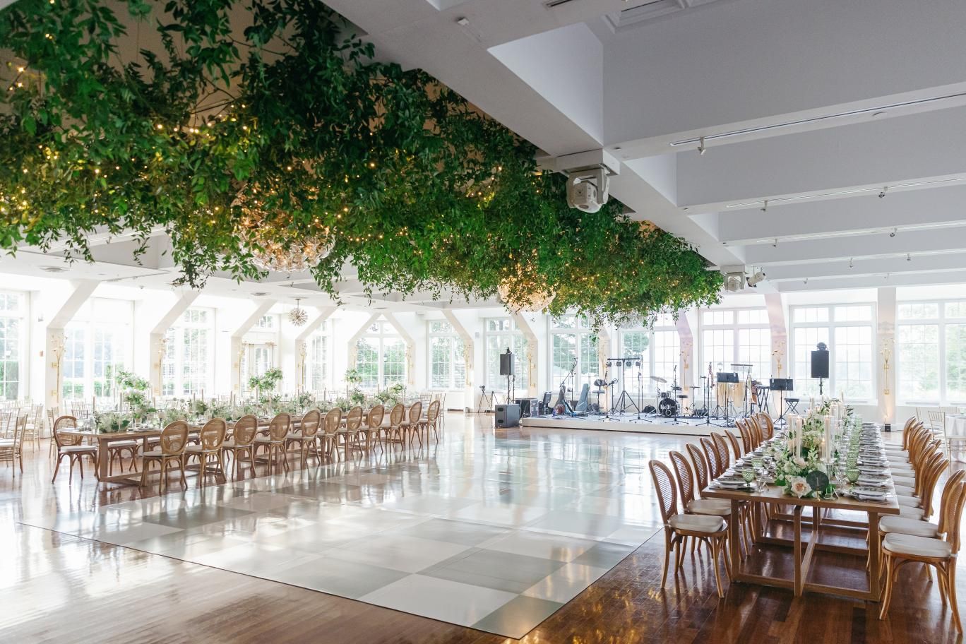 Elegant wedding reception venue with green foliage ceiling decor and wooden chairs arranged in a bright modern space.